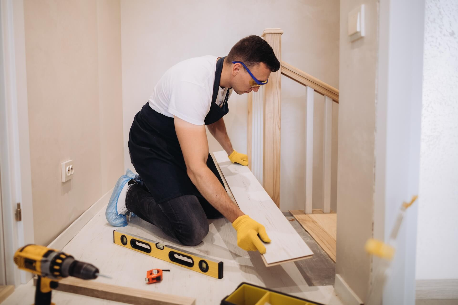 Rakentaja on polvillaan asentamassa vaaleaa lattialautaa lattialle, työkalut kuten porakone ja vatupassi ovat lähellä. Taustalla näkyy puinen portaikko, joka on osa kodin remontointia.