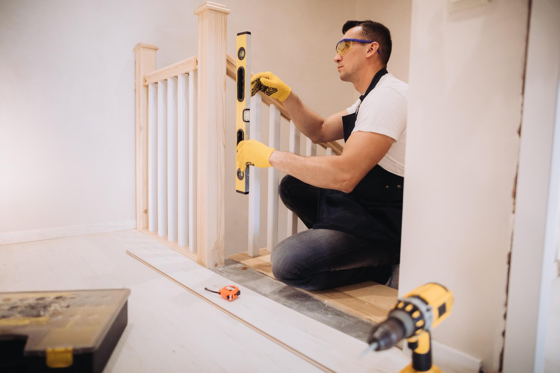 Rakentaja asentaa puisen portaan kaidetta, tarkistaen suoruutta vatupassilla. Hän on kyykyssä ja käyttää keltaisia käsineitä.